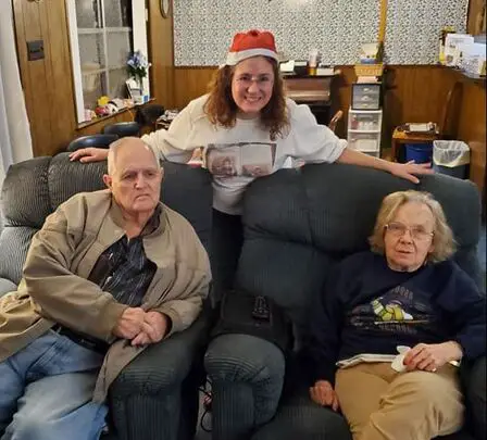 Adult daughter caregiver wearing a Santa hat leans over the backs of two chairs containing her elderly parents.